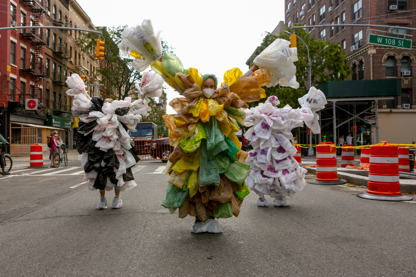 Down a New York street rimmed with traffic cones, wo women covered in white plastic bags walk behind a woman in green and yellow plastic bags. They all have their arms in a 5.